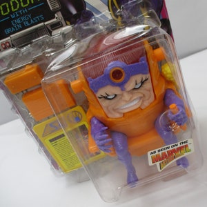 May include: A plastic action figure of the Marvel Comics character MODOK, in his signature orange and purple costume. The figure is packaged in a clear plastic blister pack with a yellow and purple card back. The card back features the text "MODOK" and "WITH ENERGY BRAIN BLASTS".