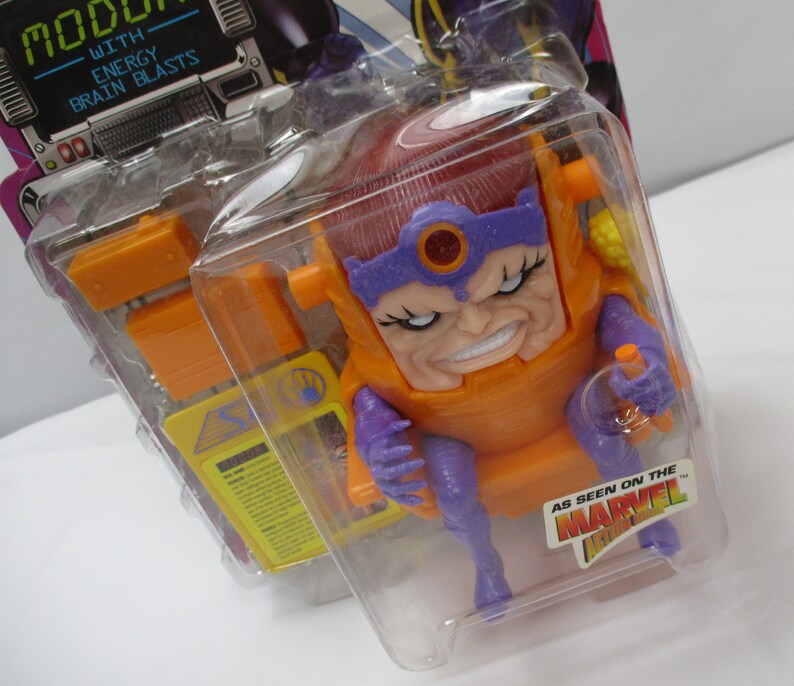 May include: A plastic action figure of the Marvel Comics character MODOK, in his signature orange and purple costume. The figure is packaged in a clear plastic blister pack with a yellow and purple card back. The card back features the text "MODOK" and "WITH ENERGY BRAIN BLASTS".