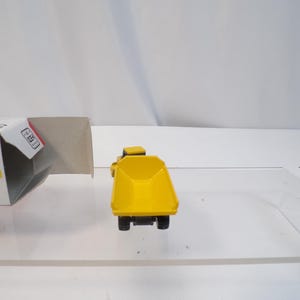 May include: A yellow toy dump truck with black wheels and a detailed design. The truck is positioned on a clear surface, with a partially visible box in the background. The toy truck is a miniature construction vehicle.