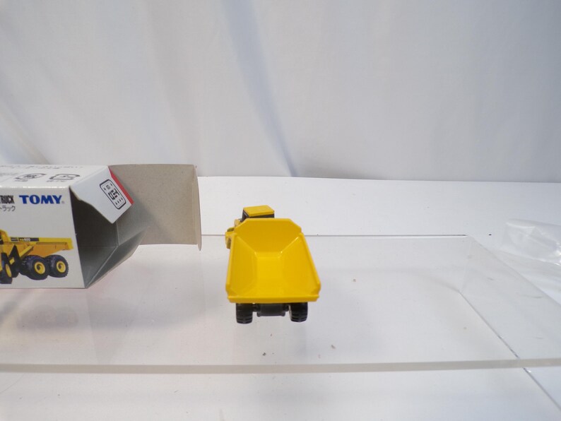 May include: A yellow toy dump truck with black wheels and a detailed design. The truck is positioned on a clear surface, with a partially visible box in the background. The toy truck is a miniature construction vehicle.