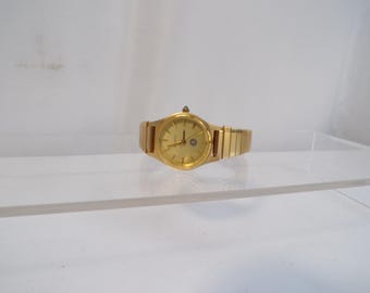 May include: A gold-toned wristwatch with a matching band. The watch face is round with gold hands and hour markers. A small diamond-like accent sits above the watch face. The band is made of rectangular links.