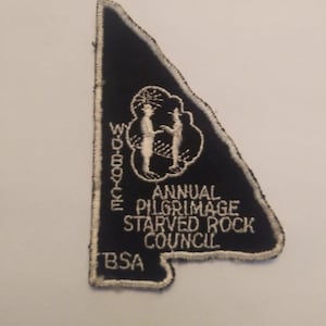 May include: A black triangular patch with white embroidered text and a design. The text reads "Annual Pilgrimage Starved Rock Council BSA" and the design depicts two figures standing in a cloud with a star above them.