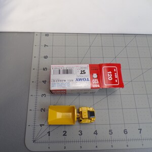 May include: A yellow toy truck with a box. The truck is made of metal and plastic. The box is red and white with Japanese text and the number 120. The truck is approximately 2 inches long.