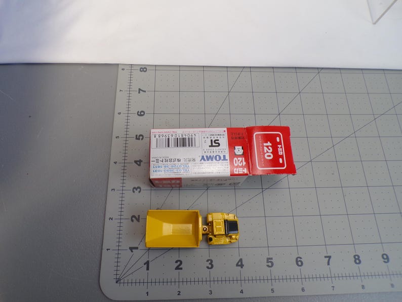 May include: A yellow toy truck with a box. The truck is made of metal and plastic. The box is red and white with Japanese text and the number 120. The truck is approximately 2 inches long.
