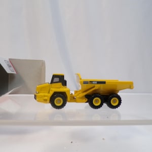 May include: A yellow toy dump truck with black wheels and a black cab. The truck is a miniature replica of a construction vehicle. The model is on a clear surface, with a partially visible box in the background.
