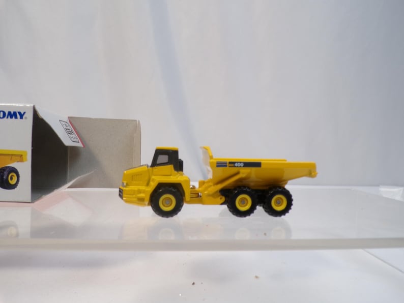May include: A yellow toy dump truck with black wheels and a black cab. The truck is a miniature replica of a construction vehicle. The model is on a clear surface, with a partially visible box in the background.