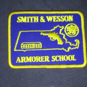 May include: Embroidered patch with a blue background and yellow border. The patch features the words "SMITH & WESSON ARMORER SCHOOL" in yellow, along with a yellow outline of a handgun and a state map.