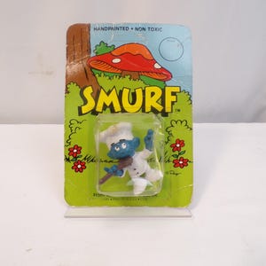 May include: A Smurf figurine wearing a chef's hat and holding a wooden spoon. The figurine is on a card with the word "SMURF" printed on it. The card is green and has a mushroom on it.