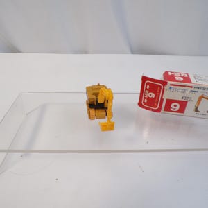 May include: A yellow and black toy excavator with a movable arm and bucket. The toy is next to its original packaging, which is white, red, and yellow. The packaging has Japanese text and an illustration of the excavator.