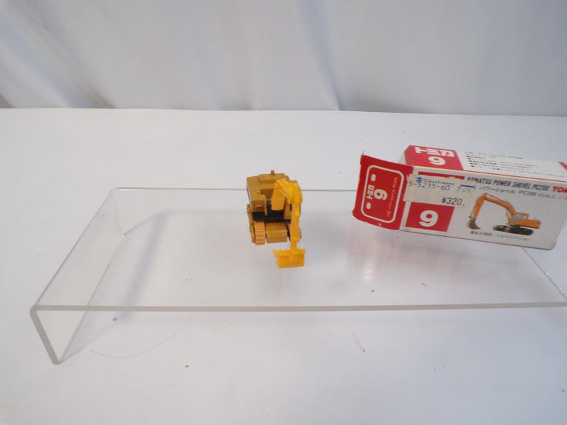 May include: A yellow and black toy excavator with a movable arm and bucket. The toy is next to its original packaging, which is white, red, and yellow. The packaging has Japanese text and an illustration of the excavator.