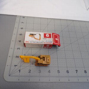 May include: A small, tan and yellow toy excavator with a matching box. The box is white and red with the number 6 on it. The toy is on a gray surface with a ruler.