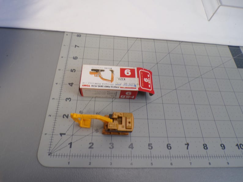 May include: A small, tan and yellow toy excavator with a matching box. The box is white and red with the number 6 on it. The toy is on a gray surface with a ruler.