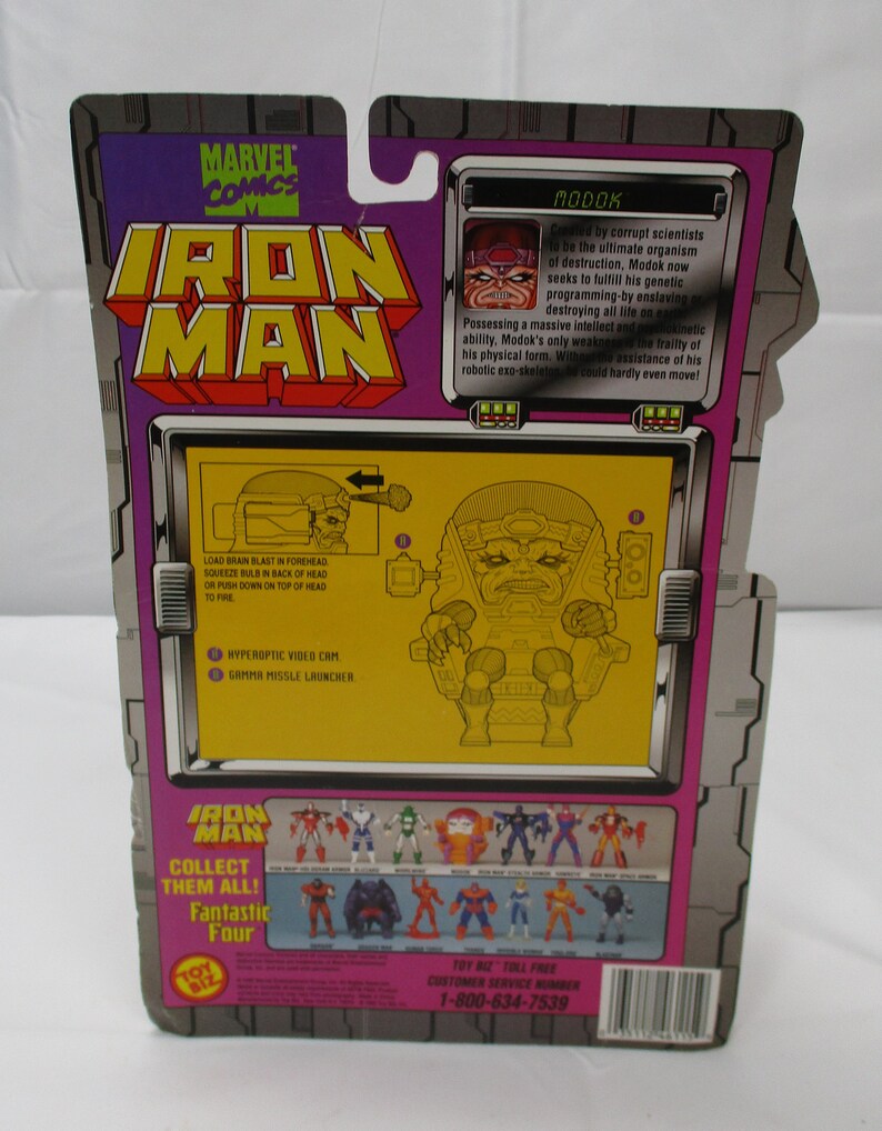 May include: A plastic action figure of the Marvel Comics character Modok, a supervillain with a large head and a robotic body. The figure is posed in a sitting position with his arms outstretched. The figure is packaged in a cardboard box with a yellow background and a picture of Modok on the back. The box also features the Marvel Comics logo and the words "Iron Man".