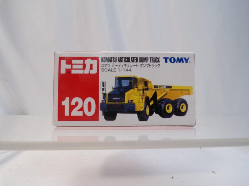 May include: A toy box featuring a yellow Komatsu articulated dump truck model. The box has the number 120 in white on a red background, with Japanese text and the TOMY logo. The truck is a scale model.