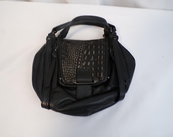 May include: Black leather handbag with a textured flap and a top handle. The bag features a buckle closure and decorative straps on the sides. The flap has a reptile-like pattern. The bag is on a white background.
