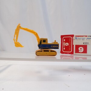 May include: A yellow and blue toy excavator with a movable arm and shovel. The toy is on a clear surface, with a red and white box in the background. The box has Japanese text and the number 9.