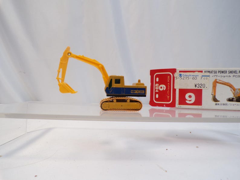 May include: A yellow and blue toy excavator with a movable arm and shovel. The toy is on a clear surface, with a red and white box in the background. The box has Japanese text and the number 9.