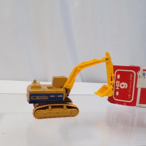 May include: A yellow and blue toy excavator with a yellow arm and bucket. The excavator is on a track system. A red and white box is behind the excavator.