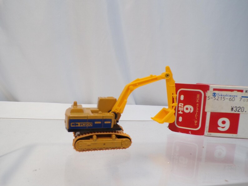 May include: A yellow and blue toy excavator with a yellow arm and bucket. The excavator is on a track system. A red and white box is behind the excavator.
