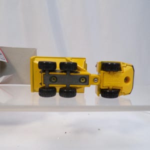 May include: Yellow toy dump truck with black wheels, viewed from underneath. The truck has a detailed design, including the undercarriage and wheel wells. The toy is on a clear surface, with a partially visible box in the background.