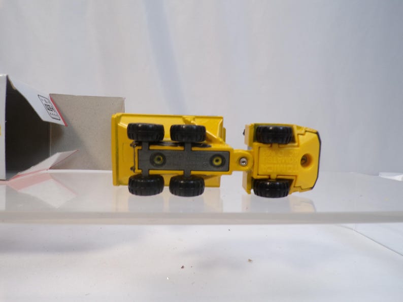 May include: Yellow toy dump truck with black wheels, viewed from underneath. The truck has a detailed design, including the undercarriage and wheel wells. The toy is on a clear surface, with a partially visible box in the background.