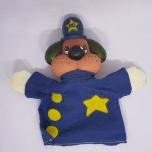 May include: A handmade dog puppet with a police officer theme. The puppet has a tan face, black nose, and green ears. It wears a blue uniform with yellow stars and circles. The hat has a star and the letters "PD".
