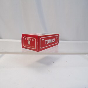 May include: A red Tomica box with white lettering and the number 9. The box is rectangular and sits on a clear surface. The Tomica logo is visible on the side of the box.