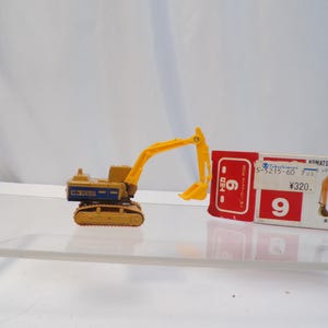May include: A yellow and tan Komatsu Power Shovel PC200 toy excavator with a blue and white label. The toy is next to a red and white box with Japanese text and the number 9.