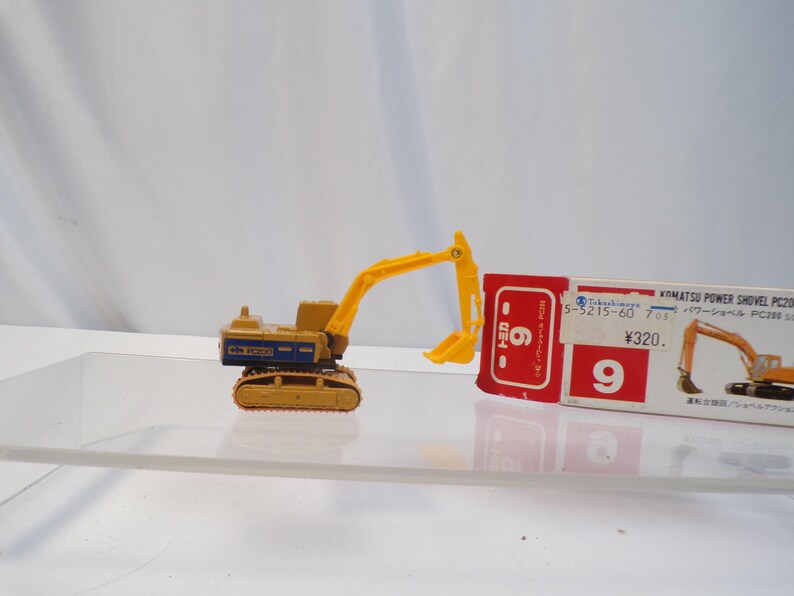 May include: A yellow and tan Komatsu Power Shovel PC200 toy excavator with a blue and white label. The toy is next to a red and white box with Japanese text and the number 9.