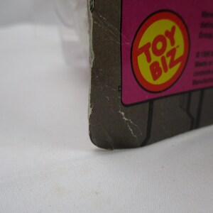 May include: Close-up of a Toy Biz logo on a pink and gray cardboard background. The logo is a red circle with a yellow center and the words "Toy Biz" in white.