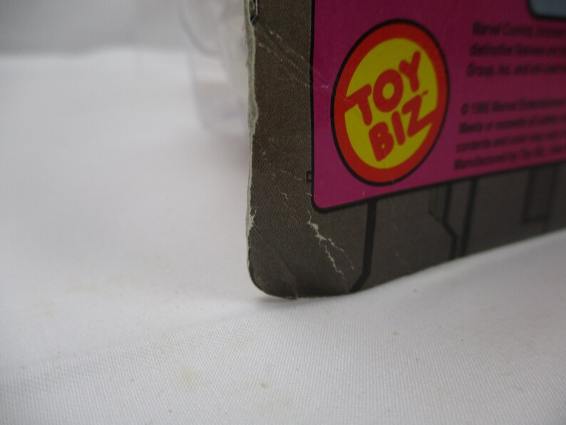 May include: Close-up of a Toy Biz logo on a pink and gray cardboard background. The logo is a red circle with a yellow center and the words "Toy Biz" in white.