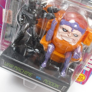 May include: A packaged action figure set featuring War Machine and M.O.D.O.K. The War Machine figure is black and silver, while M.O.D.O.K. is orange, purple, and red. The packaging includes the text "WAR MACHINE vs. M.O.D.O.K."