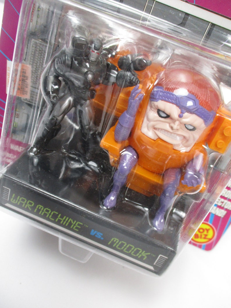 May include: A packaged action figure set featuring War Machine and M.O.D.O.K. The War Machine figure is black and silver, while M.O.D.O.K. is orange, purple, and red. The packaging includes the text "WAR MACHINE vs. M.O.D.O.K."