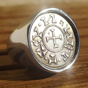 Könnte beinhalten: Ein silberner Ring mit einem Goldmünzen-Design, das ein Kreuz und andere Symbole zeigt. Die Münze ist von einem Perlenrand umgeben.