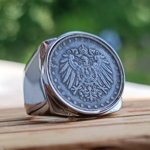 Könnte beinhalten: Ein silberner Ring mit einem detaillierten Design eines gekrönten Adlers und eines Schildes. Der Ring liegt auf einem hölzernen Untergrund.