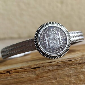 May include: A silver bracelet with a braided band and a coin charm featuring the Spanish coat of arms and the text "REY CONST. P.C. DE ESPANA 50 CENT."
