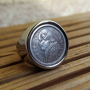 May include: Silver ring with a coin featuring a profile of Pope Pius IX. The coin is set in a silver ring band.