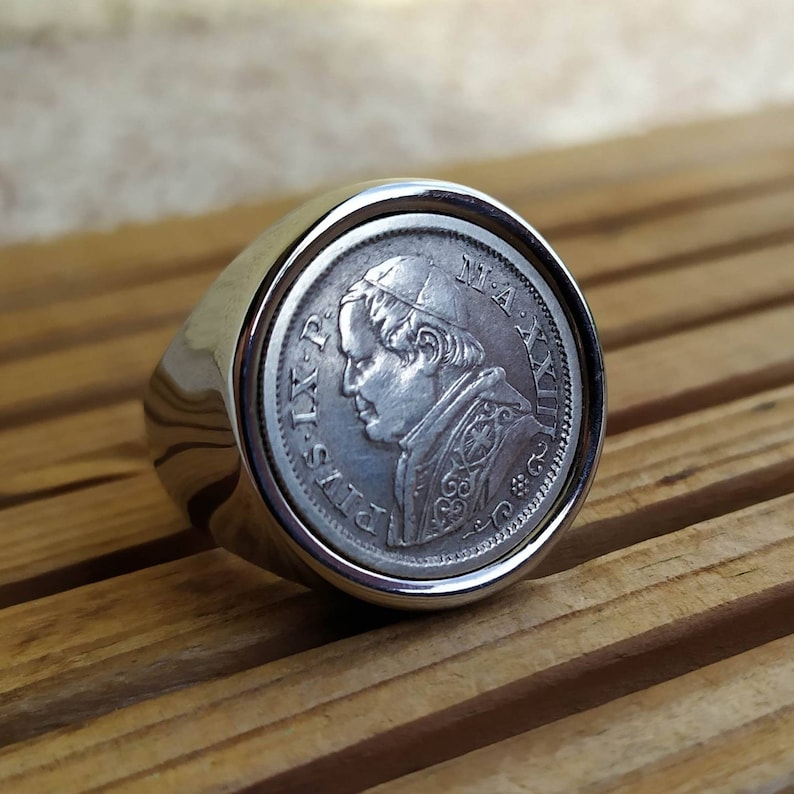 May include: Silver ring with a coin featuring a profile of Pope Pius IX. The coin is set in a silver ring band.