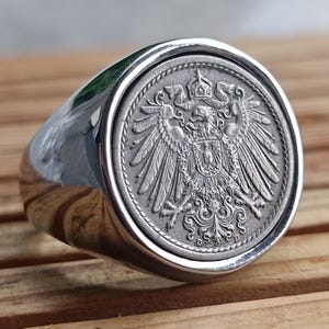 Puede incluir: Un anillo de plata con una pieza central en forma de moneda. La moneda presenta un diseño en relieve detallado de un águila con una corona y un escudo. El anillo tiene una banda pulida y redondeada y un engaste circular para la moneda.
