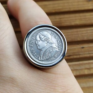 May include: A silver ring with a coin featuring a profile of Pope Pius IX. The coin is set in a silver bezel and the ring is worn on a finger.