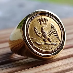 May include: A gold-colored signet ring with a detailed emblem. The emblem features an eagle with outstretched wings, a crown, and other intricate details. The ring has a polished, rounded band.