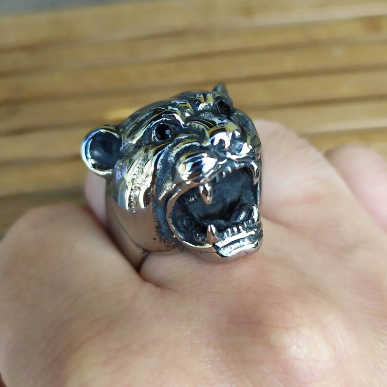 May include: A silver ring featuring a detailed design of a snarling animal head. The animal has black eyes and sharp teeth.
