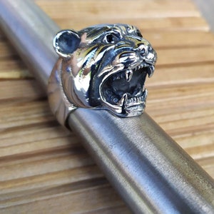 May include: A silver ring featuring a detailed, snarling tiger head design. The tiger's mouth is open, revealing sharp teeth. The ring is on a silver surface.