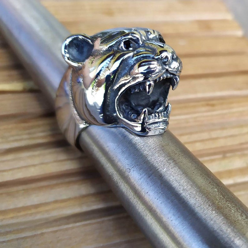 May include: A silver ring featuring a detailed, snarling tiger head design. The tiger's mouth is open, revealing sharp teeth. The ring is on a silver surface.