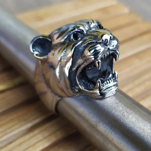 May include: A silver ring featuring a detailed, snarling panther head design. The panther's mouth is open, revealing sharp teeth. The ring is made of metal and has a polished finish.