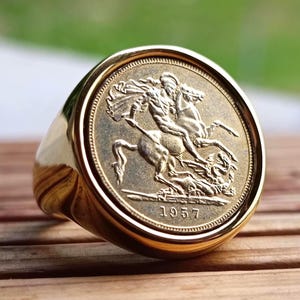 Peut inclure: Une bague dorée ornée d'une pièce de monnaie détaillée. La pièce représente une figure à cheval, transperçant un dragon, avec l'année 1957 visible. La bague a un anneau arrondi et poli et une lunette circulaire.