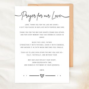 Puede incluir: Una tarjeta blanca con el texto "Prayer for our Love" en escritura elegante. La tarjeta contiene una oración con un símbolo de corazón en la parte inferior. La tarjeta está sobre un sobre marrón.