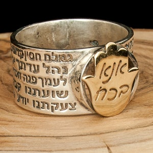 Puede incluir: Un anillo de plata con un amuleto de mano de Hamsa de oro. El anillo está grabado con texto hebreo. El anillo está sobre una superficie de madera.