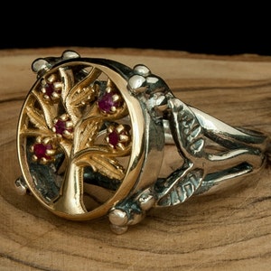 Puede incluir: Un anillo de plata con un dise&ntilde;o de &aacute;rbol de la vida en oro y piedras preciosas rojas. El anillo est&aacute; sobre una superficie de madera.