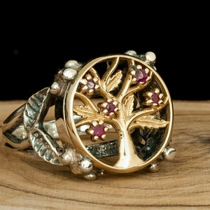 Puede incluir: Un anillo de plata con un dise&ntilde;o de &aacute;rbol de la vida en oro y piedras de rub&iacute; rojas. El anillo est&aacute; colocado sobre una superficie de madera.